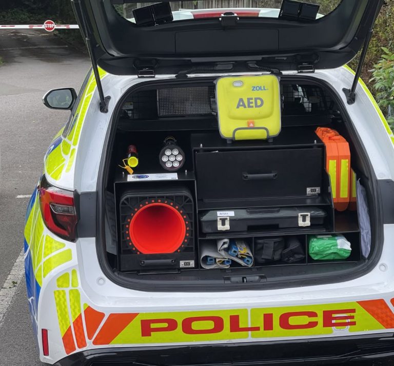 The open boot of a white police car featuring high-visibility yellow and blue Battenburg markings. A black modular storage unit fills the space, meticulously organized with emergency equipment. A bright yellow ZOLL AED (defibrillator) bag sits prominently on the top shelf. Other visible gear includes a large collapsible orange traffic cone, a heavy-duty flashlight, a fire safety stick, an orange hard case, and a green first aid kit. The rear bumper features red "POLICE" text on a reflective yellow and orange background. A barrier gate with a "STOP" sign is visible in the background.